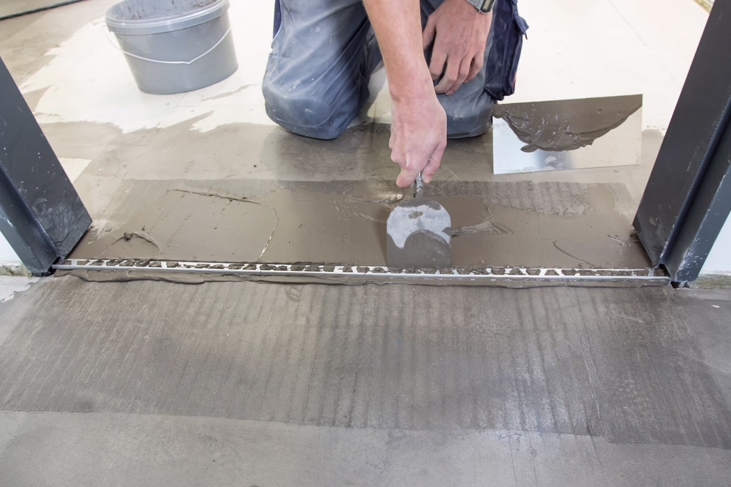 UZIN_Application Image_UZIN NC182_2018-03 A construction worker kneels at an interior doorway, applying grey tile adhesive or floor leveling compound with a notched trowel. He spreads and smooths the wet material over a metal transition profile at the threshold; a second trowel with compound and a grey bucket are visible nearby. The image focuses on the worker's hands and lower torso clad in work trousers with paint specks, showing the ridged adhesive bed in the foreground, fresh smoothing marks and the careful alignment of two floor surfaces.