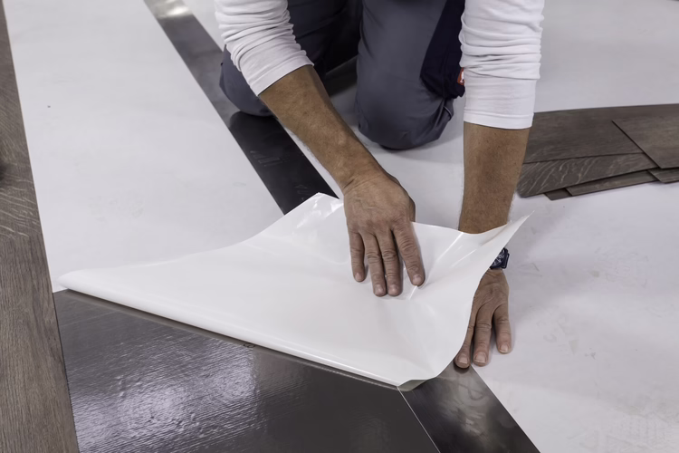 Mark the starting point of the first row of planks or tiles with a suitable pen on the protective paper. Place a steel rail along the measuring points and align. Pull the protective paper away from the Sigan Elements Plus and tear off the protective paper along the steel rail. The straight line thus creates a reference line of the first row of planks or tiles.